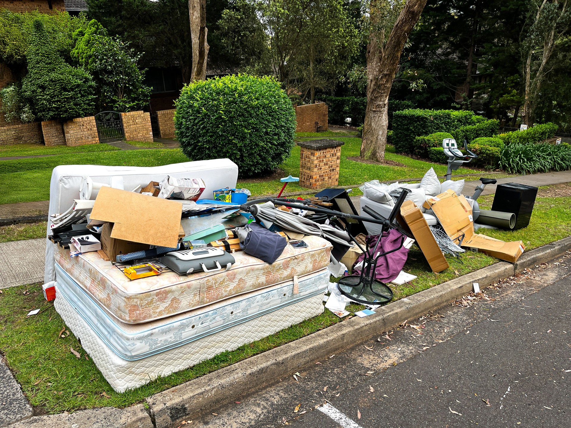 A pile of rubbish on the curb for council collection, including cardboard boxes, a couch with pillows, stacked mattresses, and coat hangers